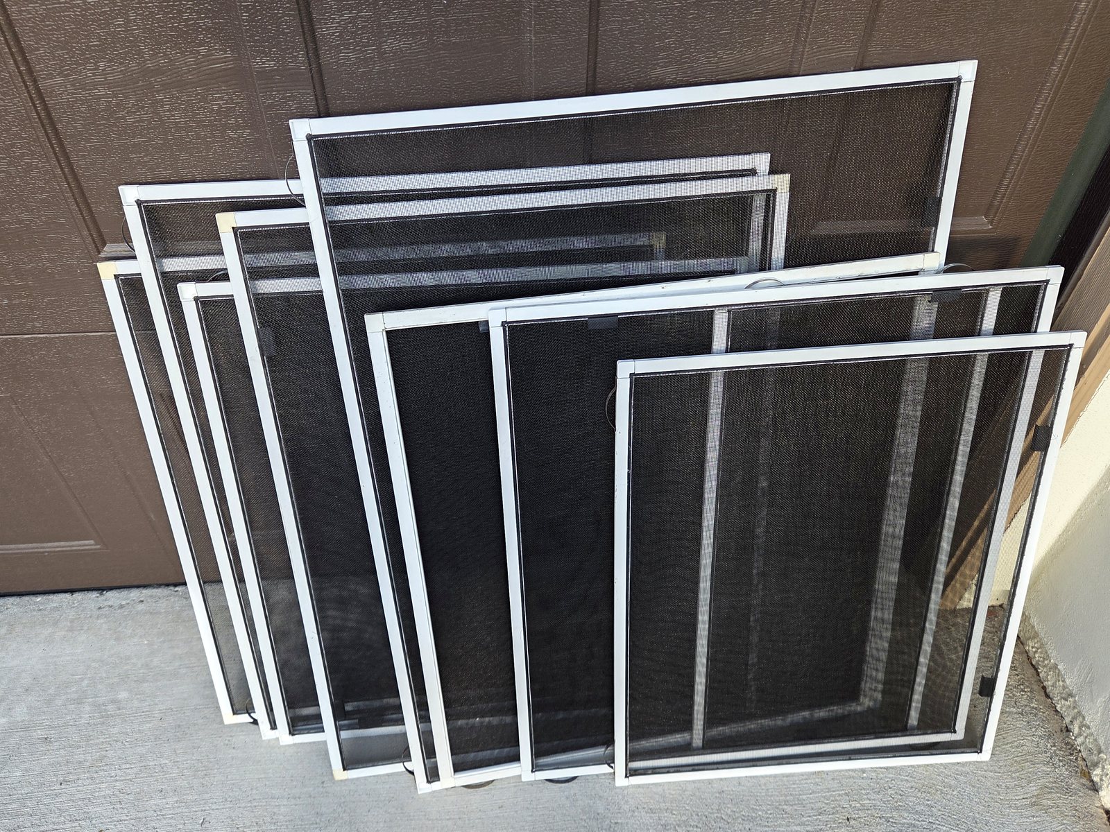 Completed batch of white-framed window screens with charcoal mesh arranged in front of a garage