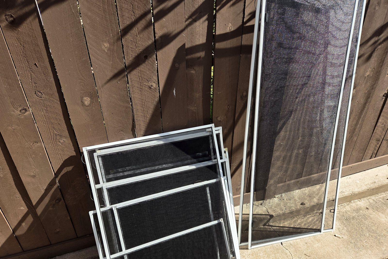 Completed white-framed window screens of several sizes leaning against a brown fence